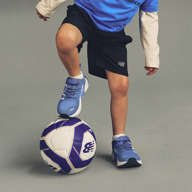 One foot is placed on top of a white and purple soccer ball with a visible New Balance logo, while the other foot stands on the floor. 