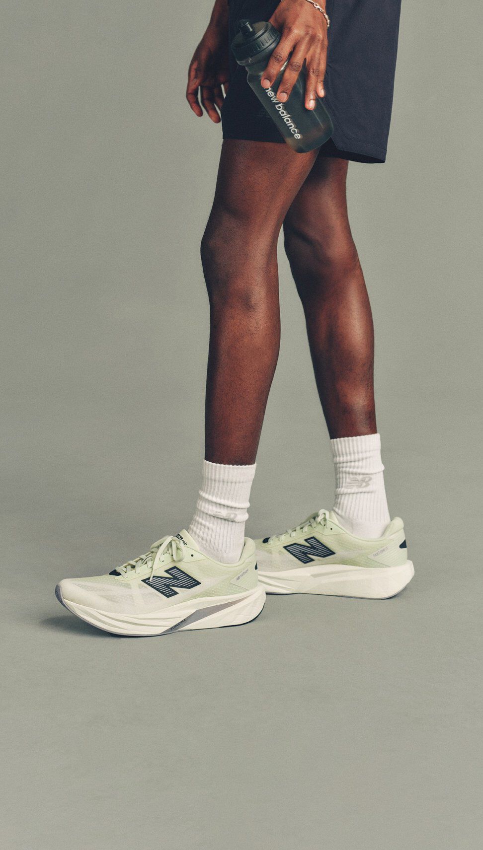 Close-up studio image of light green New Balance running shoes paired with white crew socks and black shorts, with a hand holding a water bottle at the side against a neutral gray background.