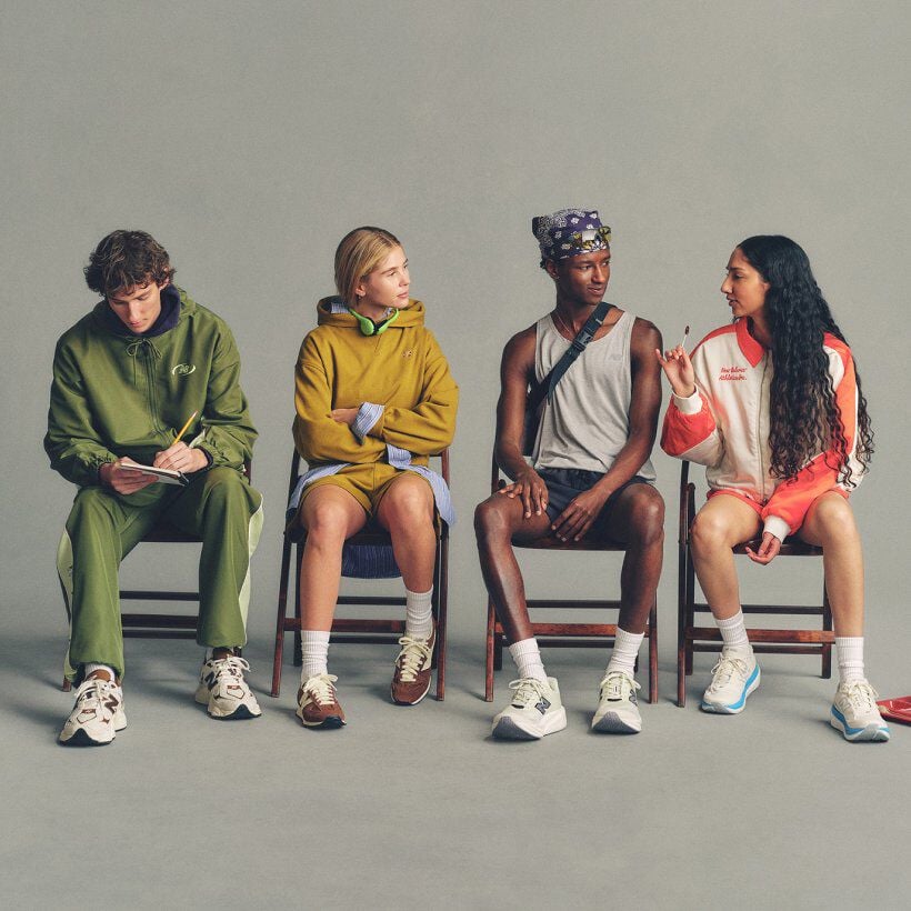 Four people seated in a row on chairs against a plain background, each wearing casual outfits and New Balance sneakers, with one writing in a notebook and others conversing.