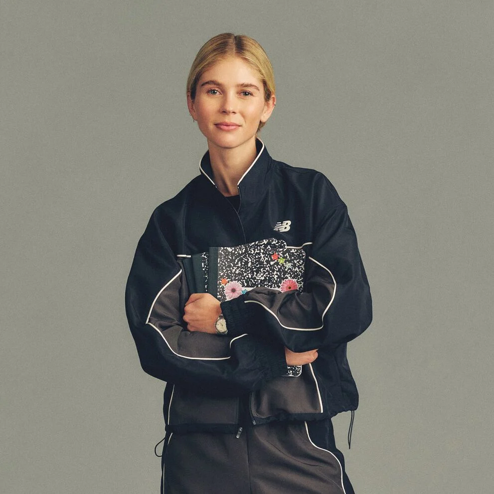 The person is wearing a black track jacket with light piping details and a small New Balance logo on the chest, paired with matching black pants.