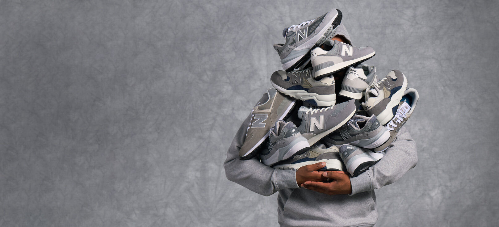A person wearing a grey New Balance hoodie, holding a pile of grey New Balance shoes in their arms with their face obscured.