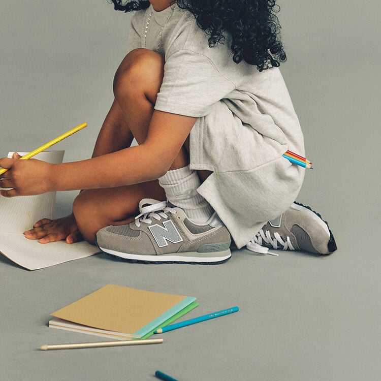 Close-up studio image of a child crouching on the floor against a plain light gray background, focused on drawing on a sheet of lined paper. 