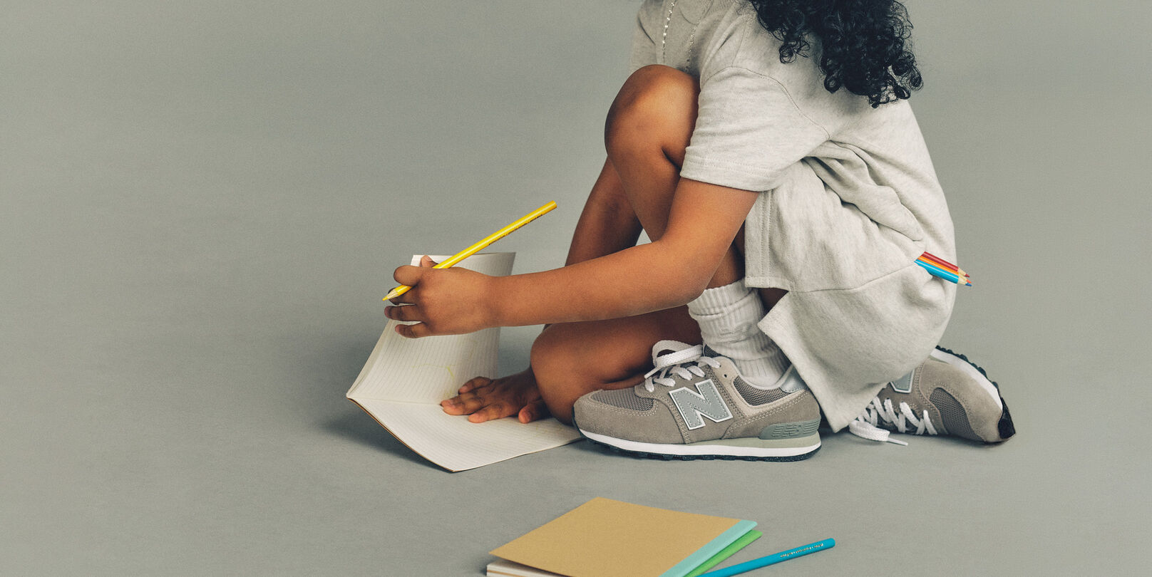 Close-up studio image of a child crouching on the floor against a plain light gray background, focused on drawing on a sheet of lined paper. 