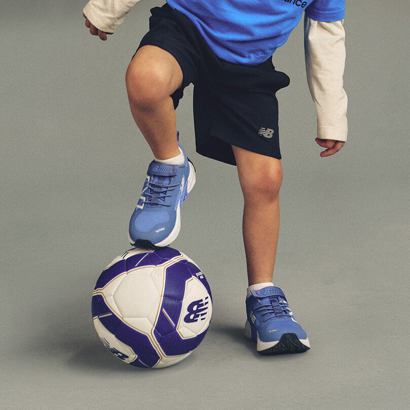 One foot is placed on top of a white and purple soccer ball with a visible New Balance logo, while the other foot stands on the floor. 