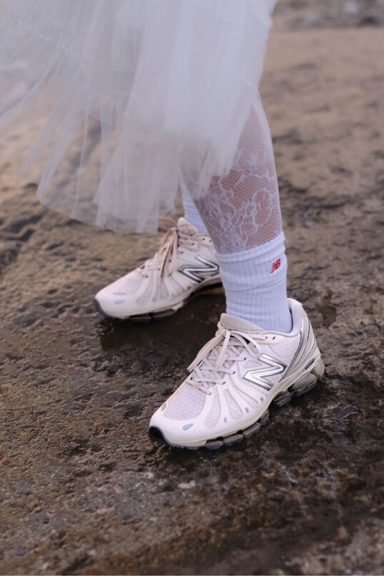 Close-up of someone wearing white New Balance sneakers on a gravel background.