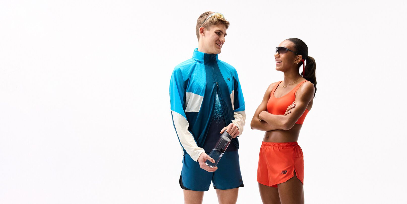 Two people in colorful athletic outfits stand facing each other on a plain white background, one holding a water bottle, both wearing sporty accessories and appearing mid-conversation.