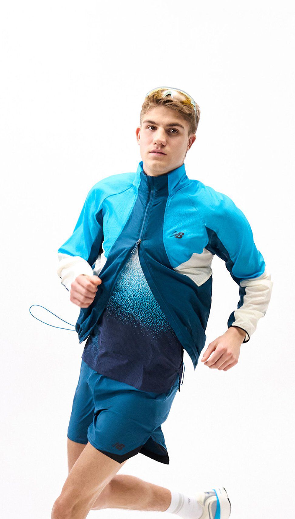 Person running in a blue color blocked jacket and teal shorts, mid-stride against a clean white background.